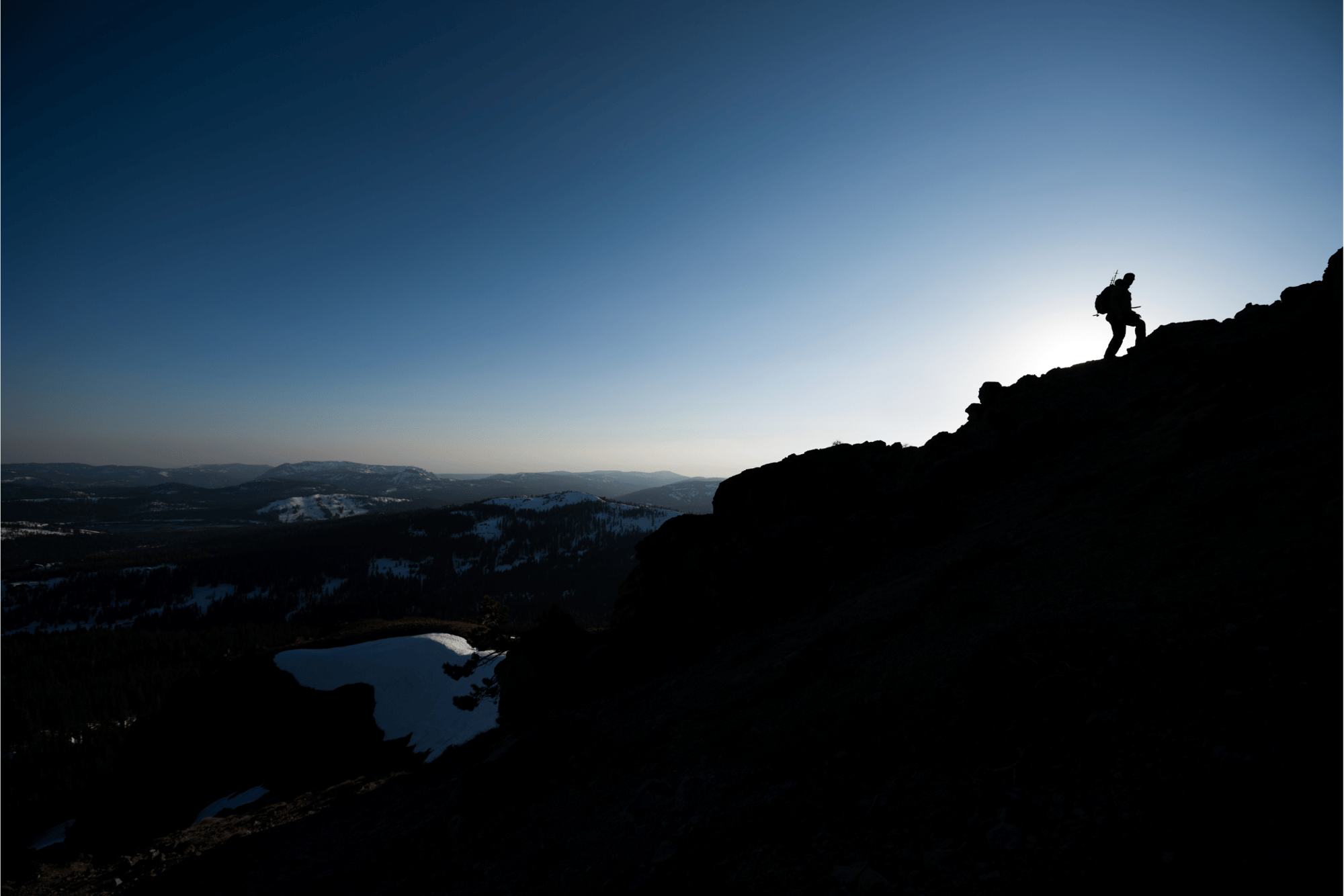 Silueta de un excursionista en la cima de una montaña al amanecer, con vistas a un paisaje montañoso y un lago parcialmente congelado. El cielo sobre el horizonte es claro y azul.