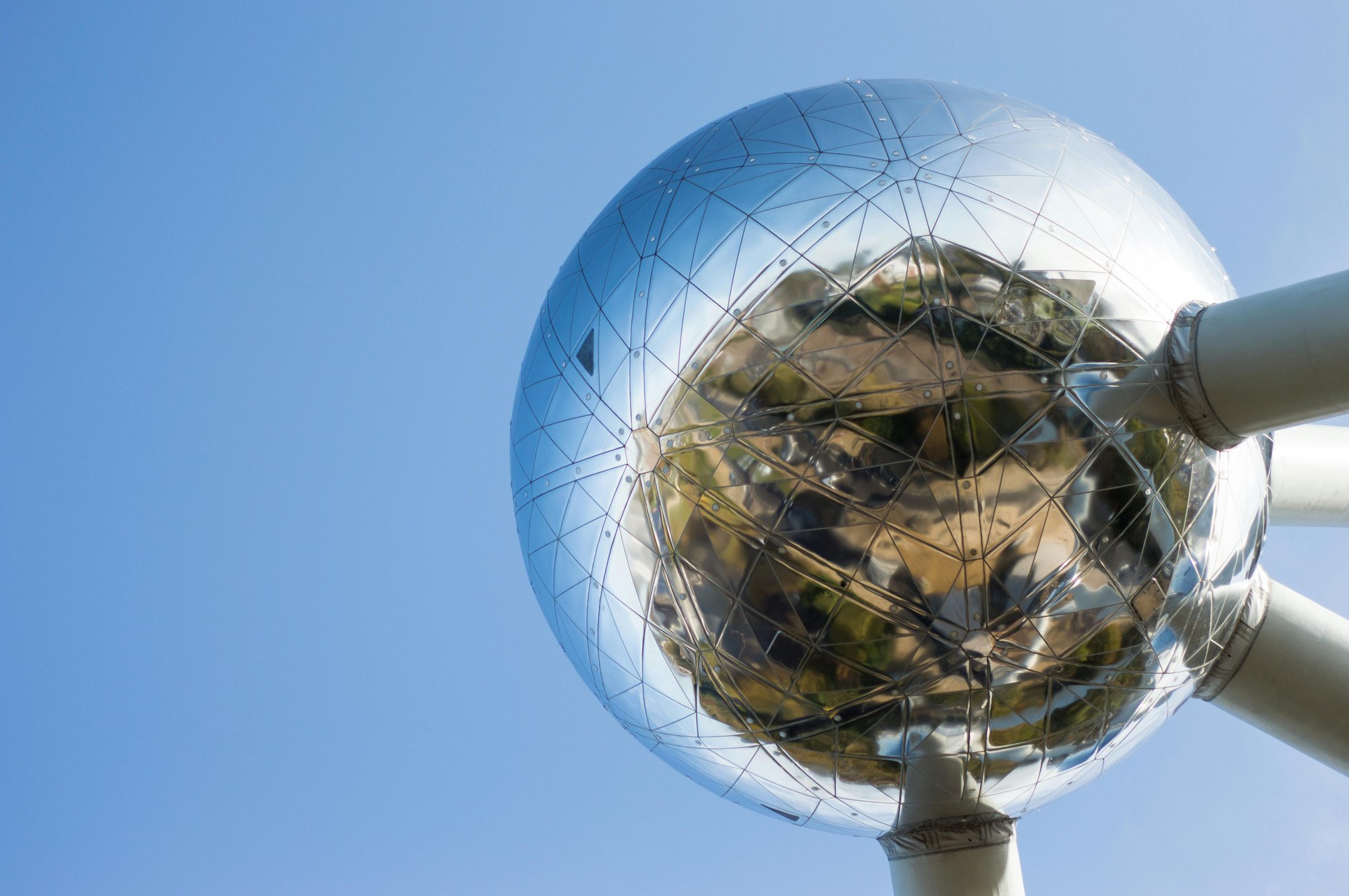 Primer plano de una esfera de acero del Atomium, mostrando su superficie metálica y geométrica con reflejos de árboles y el cielo azul.